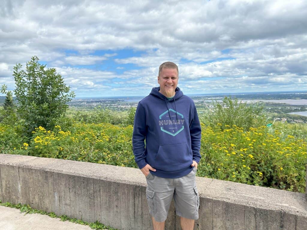 A man with a beard, wearing a blue hoodie and gray shorts, stands by a low stone wall with a background of green bushes and yellow flowers. Behind him, there is a clear view of the cityscape and an expansive cloudy sky.